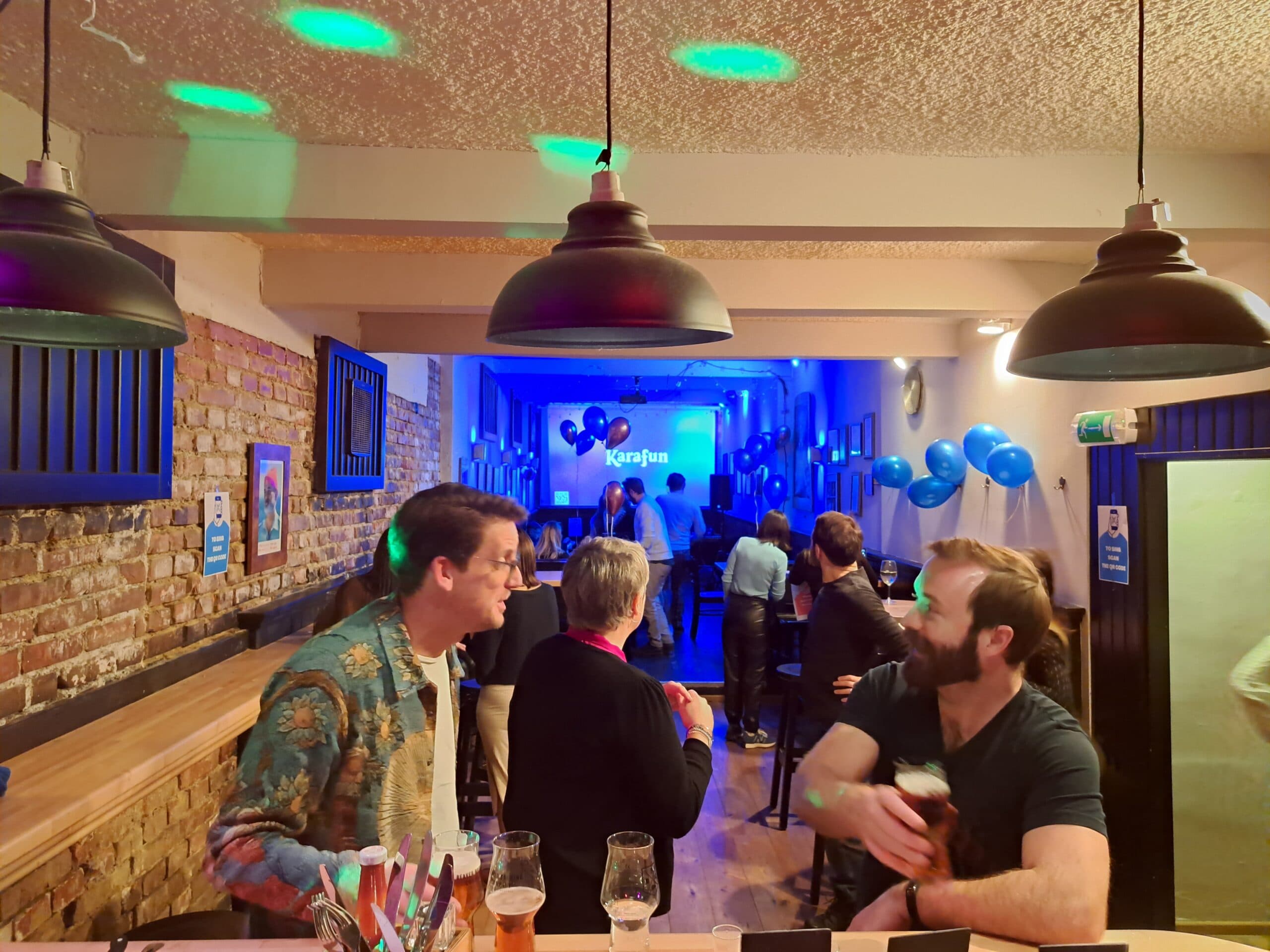Feier im Partyraum: Karaoke-Leinwand, blaue Luftballons, volle Tische